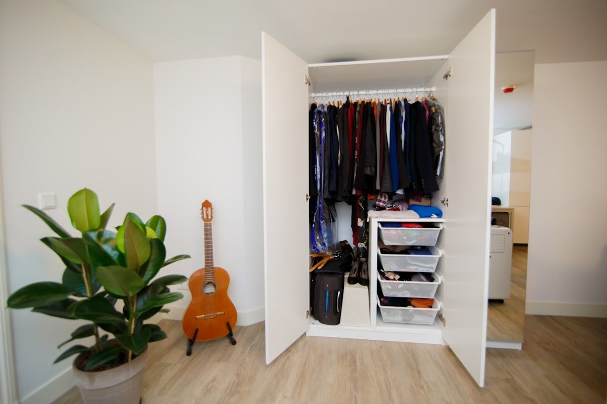 A crowded shared bedroom closet with one side packed with hanging clothes, shoes spilling onto the floor, a chair covered in sweaters, and a blocked full-length mirror near a narrow opening where two wardrobes are overlapping and creating clutter.