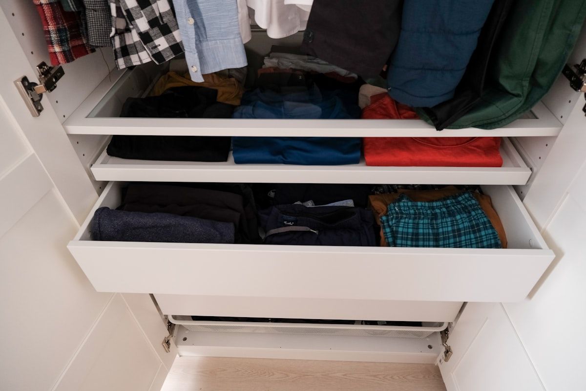 A bedroom closet with a rod, shelves, and drawers arranged around everyday use: shirts and pants hanging at eye level, shoes lined neatly on the floor, folded sweaters on a middle shelf, and a few storage bins on the top shelf, with one chair nearby holding a stray pile of clothes to show the contrast between organized and disorganized storage.