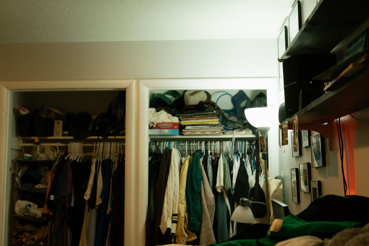 The floor of a small closet with a low-profile shoe rack holding several pairs in a tidy row beneath hanging clothes, alongside a tall narrow shoe organizer and a few off-season shoes stored in a labeled bin on a higher shelf.